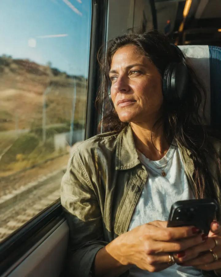 A woman in her fifties sits alone by a train window, headphones on, her phone resting in her lap.