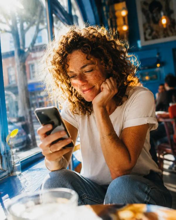A woman in her early thirties sits by a café window, phone in hand.