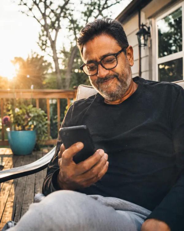 A man in his fifties sits on a porch in late afternoon light, looking at his phone.